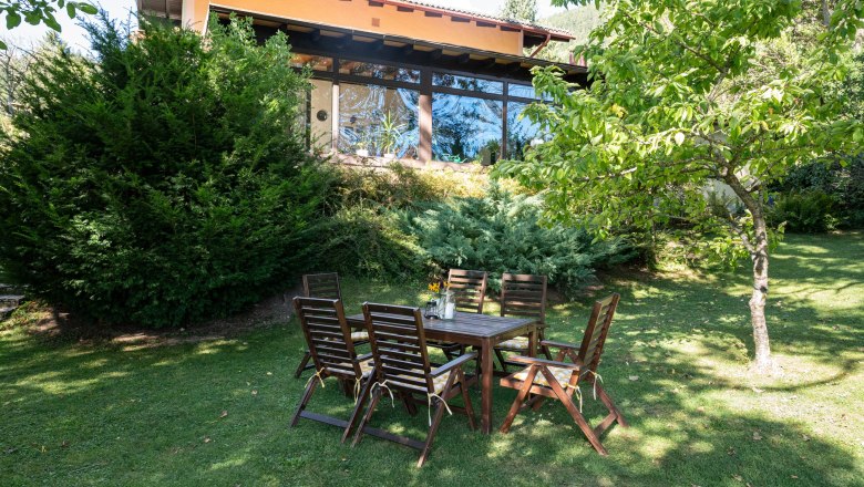 Garten, Ferienhaus Schlager, © Claudia Schlager Ein Garten mit Holztisch und Stühlen vor einem Ferienhaus mit großen Fenstern und umgeben von Bäumen und Sträuchern.