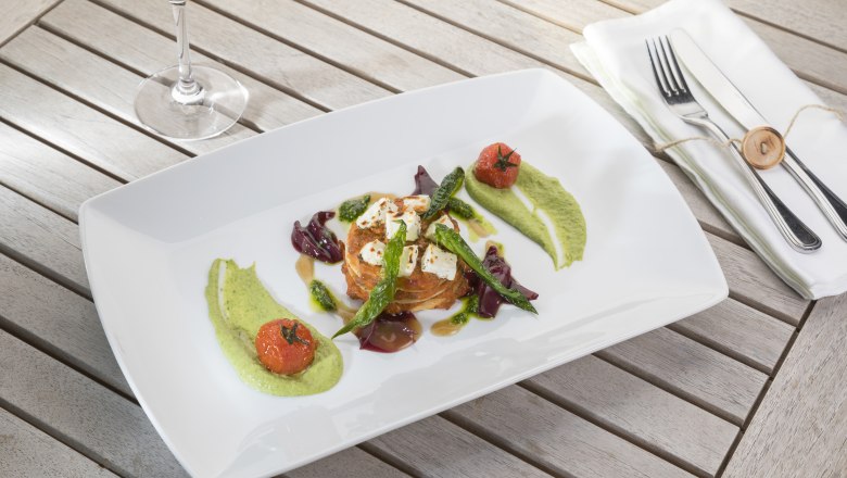 The heart of the business: fresh cuisine with a biker-friendly touch, © Niederösterreich Werbung/Kurt Pinter Gourmet dish on a white plate with vegetables and sauces, accompanied by a glass of white wine.