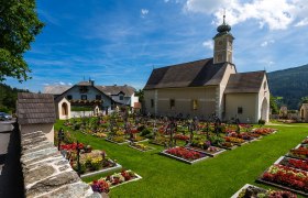 Pfarrkirche St. Peter am Neuwald, © Wiener Alpen, Kremsl Pfarrkirche St. Peter am Neuwald mit gepflegtem Friedhof und blühenden Blumenbeeten, umgeben von grüner Landschaft und blauem Himmel.