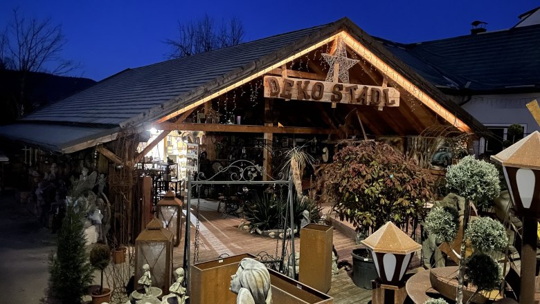 Dekostadl Pernitz, © Wiener Alpen/Katharina Lechner Ein dekorativ beleuchtetes Holzgebäude mit der Aufschrift 'Deko Stadl' bei Nacht, umgeben von Pflanzen und Dekorationsartikeln.