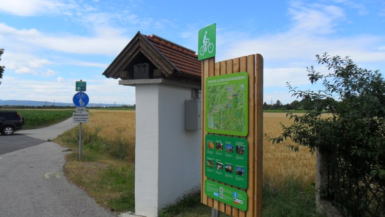 Radstartplatz-Tafel Katzelsdorf, © ©Wiener Alpen Radstartplatz-Tafel in Katzelsdorf mit Wegweiser und Landschaft im Hintergrund.