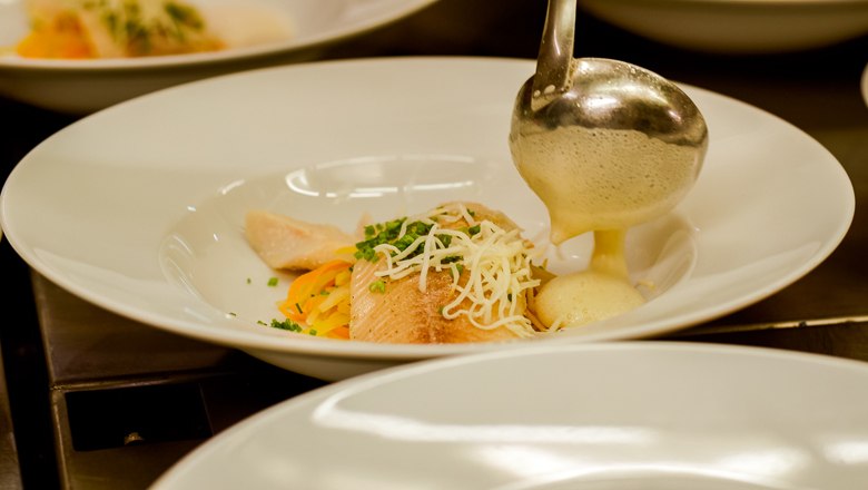 6-gängiges Hauben-Menü, © Wiener Alpen/Kremsl Ein Teller mit Fisch, Gemüse und einer hellen Soße, die mit einer Kelle serviert wird.