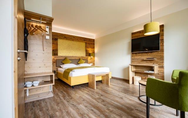 Das Gästehaus im Triad, © Marketing Platzhirsch, Gobauer Modernes Hotelzimmer mit Holzmöbeln, Doppelbett, grünem Sessel und Flachbildfernseher.