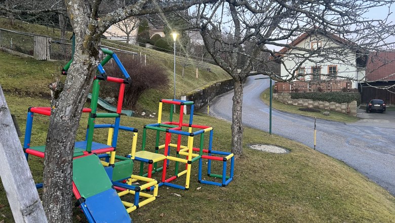 Spielplatz, © Andrea Tauchner Bunter Kletterspielplatz mit Rutsche auf einer Wiese neben einer Straße und einem Haus.