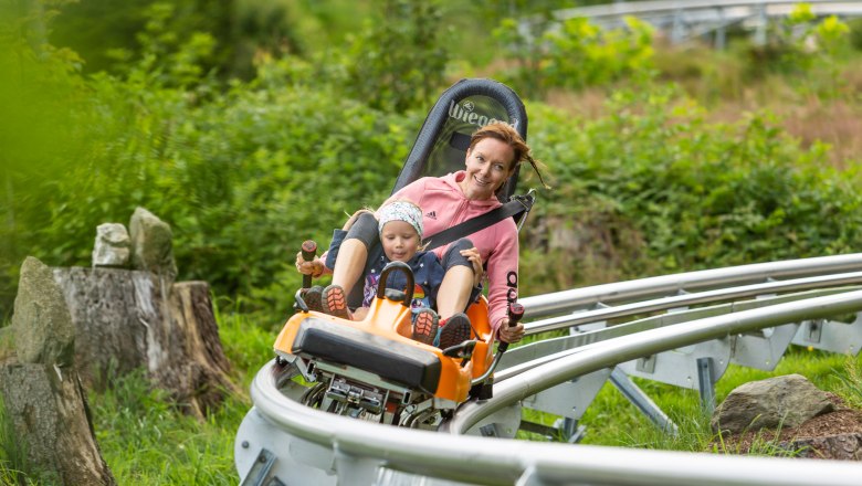 Sommerrodelbahn, © Erlebnisarena St. Corona Eine Frau und ein Kind fahren auf einer Sommerrodelbahn durch eine grüne Landschaft.