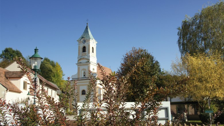 Pfarrkirche Walpersbach, © Roffeis Heinz Pfarrkirche Walpersbach mit Kirchturm und umliegenden Gebäuden, umgeben von Bäumen und Sträuchern unter blauem Himmel.