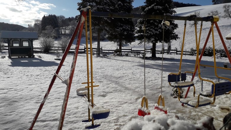 Garten der Pension Hendling, © Josef Hendling Schneebedeckter Spielplatz mit Schaukeln und Bäumen im Hintergrund.