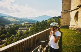 Pause bei Wolfgangskirche in Kirchberg am Wechsel, © Wiener Alpen, Schönauer Eine Radfahrerin mit Helm steht vor einer alten Kirche, im Hintergrund der Ort Kirchberg am Wechsel und Hügel