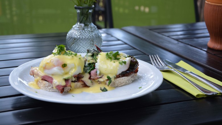Schlemmen im Turmcafé, © dasTurmcafé - Gemeinde Scharzenbach Ein Teller mit Eggs Benedict auf einem Holztisch, daneben Besteck und eine Vase mit Blumen.