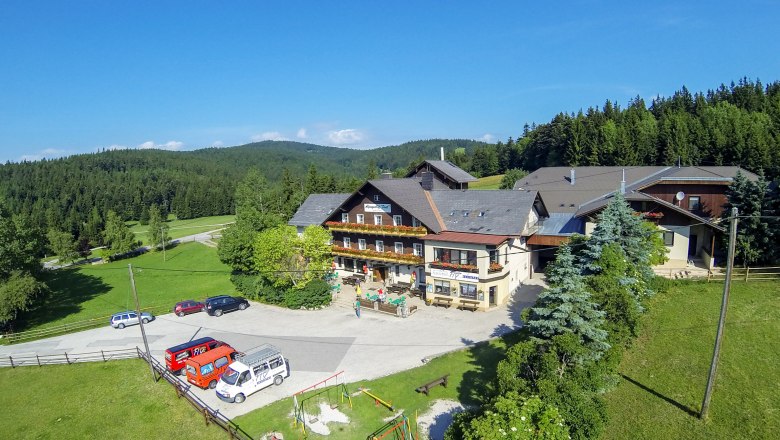 Alpengasthof Postl, © Wiener Alpen, Foto: Franz Zwickl Alpengasthof Postl in einer grünen, bewaldeten Landschaft mit mehreren geparkten Autos und einem Spielplatz im Vordergrund.