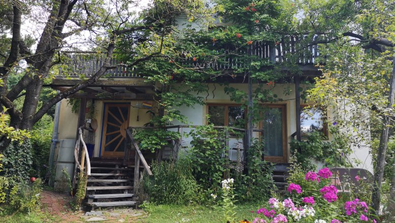 Waldgarten an der Rosalia, © "Natur im Garten" Ein Haus mit Holzbalkon, umgeben von Bäumen und bunten Blumen im Garten.