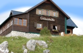 Geländehütte, © Gelaendehuette, Foto Fam. Postl Eine Berghütte mit der Aufschrift 'Geländehütte' auf einer Wiese mit Felsen und einer Kuh im Vordergrund.