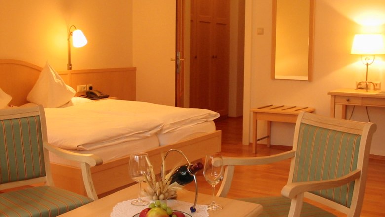 Zimmer im Hotel Belvedere, © Hotel Belvedere Ein gemütliches Hotelzimmer mit Doppelbett, zwei Stühlen, einem Tisch mit Obst und Wein, und warmem Licht.