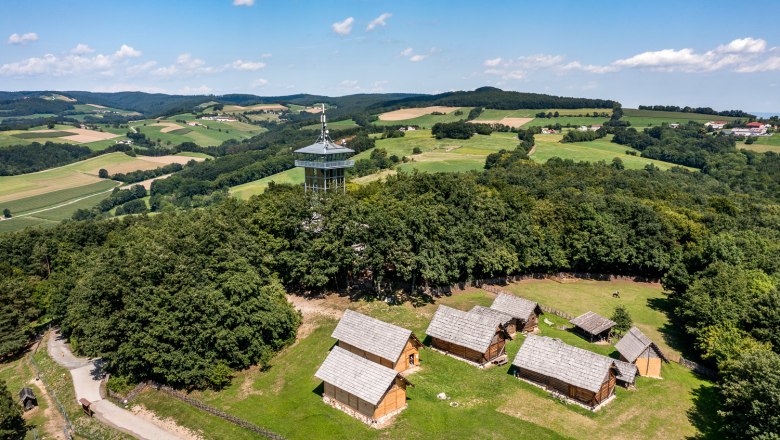 Celtic village Schwarzenbach, © Wiener Alpen, Christian Kremsl Aerial view of the Celtic village of Schwarzenbach with wooden houses and a lookout tower, surrounded by green fields and forests.