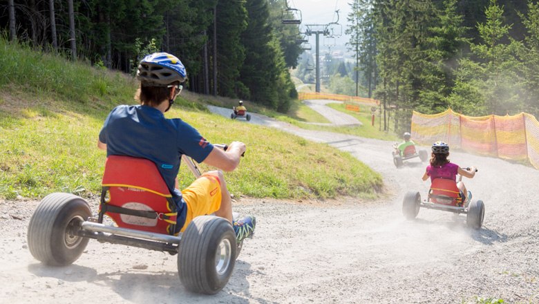 Mountaincart-Spaß in Mönichkirchen, © Erlebnisalm Mönichkirchen/Fotograf und Fee Personen fahren mit Mountaincarts einen Schotterweg hinunter, umgeben von Bäumen und einem Skilift.