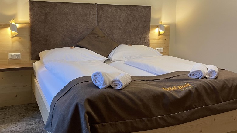 Doppelzimmer, © Hotel Post Hönig Ein gemütliches Doppelzimmer mit großem Bett, dekoriert mit brauner Bettwäsche und Handtüchern. An der Wand sind geometrische Muster.