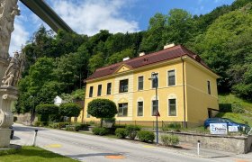 Apartment Palma, © Wiener Alpen Gelbes Gebäude mit rotem Dach neben einer Straße, umgeben von Bäumen und einer Statue im Vordergrund.