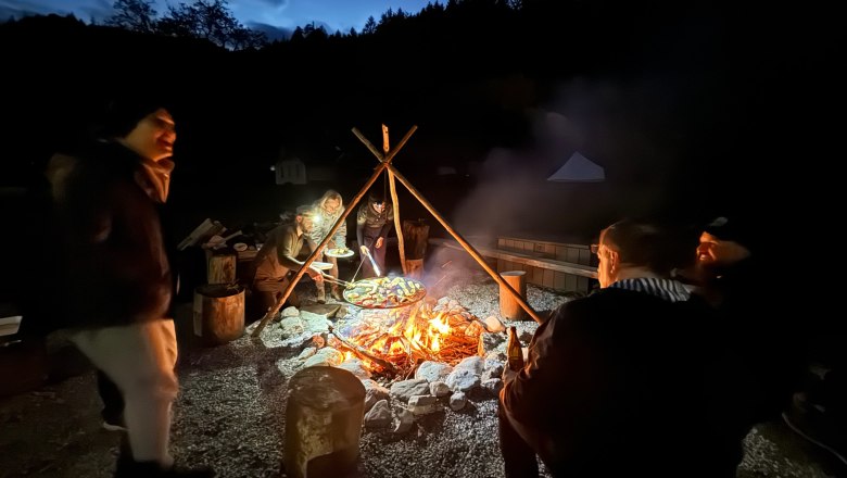 Survival Workshop Wildnis Zentrum Nasswald, © Georg Bergthaler Menschen sitzen nachts um ein Lagerfeuer im Freien, umgeben von Bäumen.
