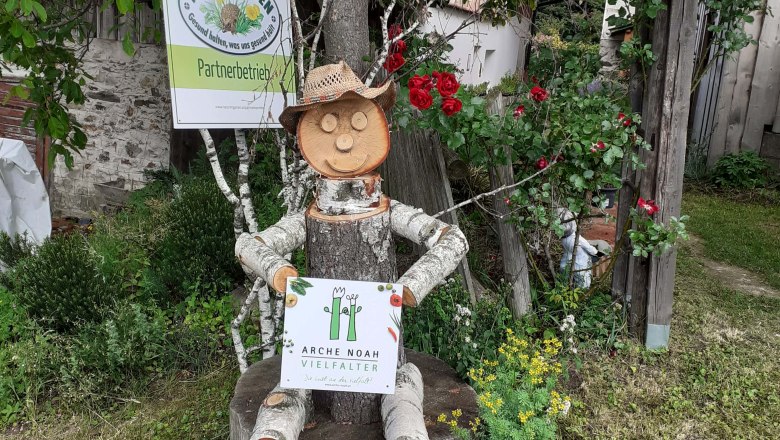 Natural garden paradise Beisteiner, © Naturgartenparadies Beisteiner Wooden figure with straw hat sitting on tree stump, holding sign with inscription 'Arche Noah Vielfalter'. Background: Nature in the garden sign