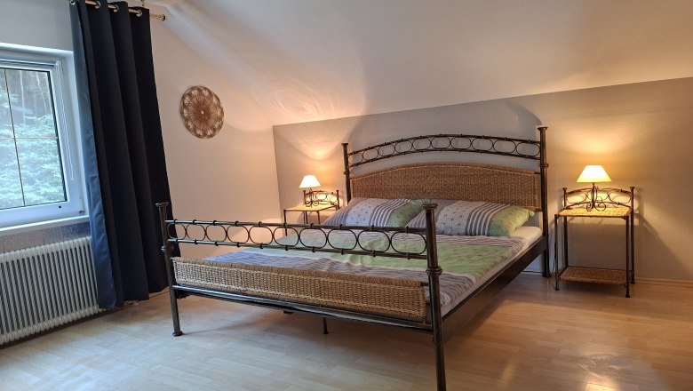 Schlafzimmer, © Manuela Heher Ein gemütliches Schlafzimmer mit einem Metallbett, zwei Nachttischen mit Lampen und einem Fenster mit dunklen Vorhängen.