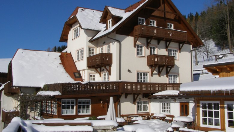 Der Althammerhof im Winterkleid, © Althammerhof, Julius Schneidhofer Ein großes, traditionelles Haus im Schnee mit Holzverzierungen und blauen Himmel im Hintergrund.