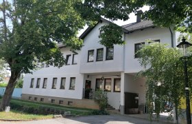 Hausansicht Heurigen Leeb, © Wiener Alpen Außenansicht eines weißen Gebäudes mit vielen Fenstern, umgeben von Bäumen.