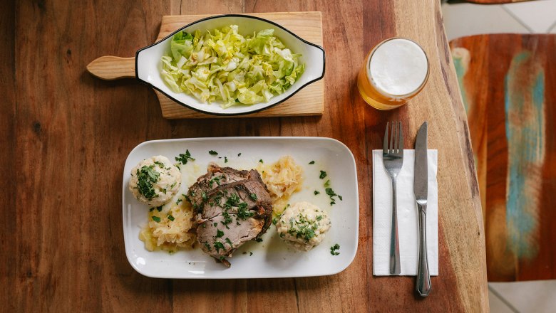 Siegl's am Markt "originalgetreu - bodenständig - ehrlich", © Wiener Alpen/Sophie Menegaldo Ein Teller mit Schweinebraten, Knödeln und Sauerkraut, daneben ein Salat und ein Glas Bier auf einem Holztisch.