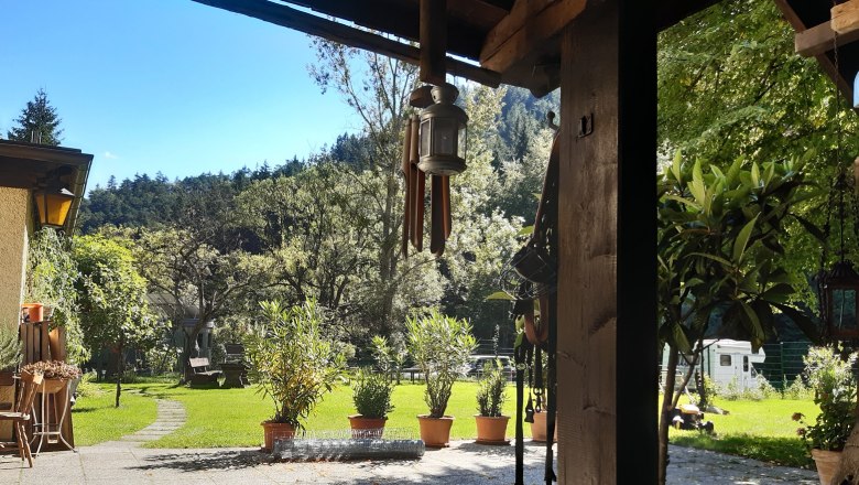 Innenhof, © Familie Kellner Ein sonniger Innenhof mit Rasen, Bäumen und Topfpflanzen. Im Vordergrund eine überdachte Terrasse mit einer Laterne und Gartenmöbeln.