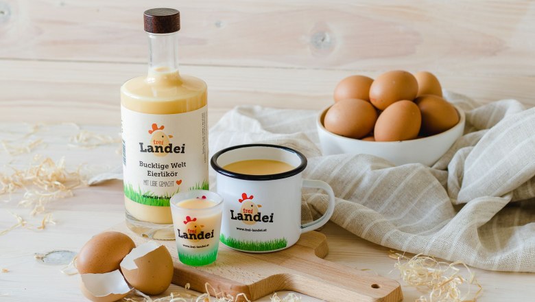 Egg liqueur, © Vikografik/ Viktoria Kornfeld A bottle, a glass and a cup filled with egg liqueur on a wooden board. In the background, a bowl of eggs on a gray cloth.