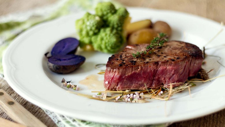 Delicious dishes from the country inn kitchen, © Sooo gut schmeckt/Viktoria Kornfeld A piece of steak on a white round plate with vegetables, potatoes and herbs.