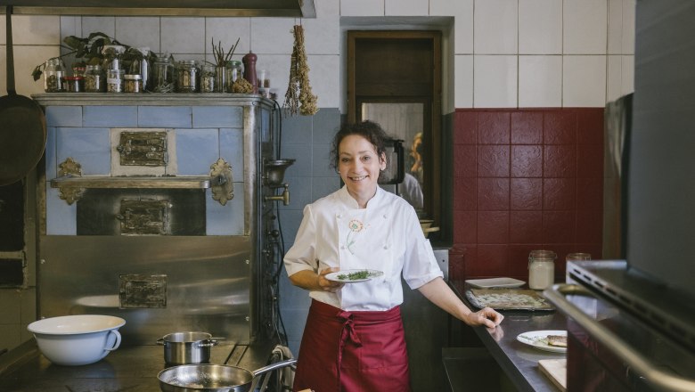 Kräuterwirtin Gerda Stocker, © Wiener Alpen/Sophie Menegaldo Eine Frau in einer Küche steht vor einem alten Herd und hält einen Teller mit Kräutern.
