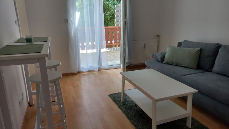 Wohnzimmer, © Dekic-Ouaja Modernes Wohnzimmer mit grauem Sofa, weißem Couchtisch und Barhockern. Balkon mit Glas-Schiebetür im Hintergrund.