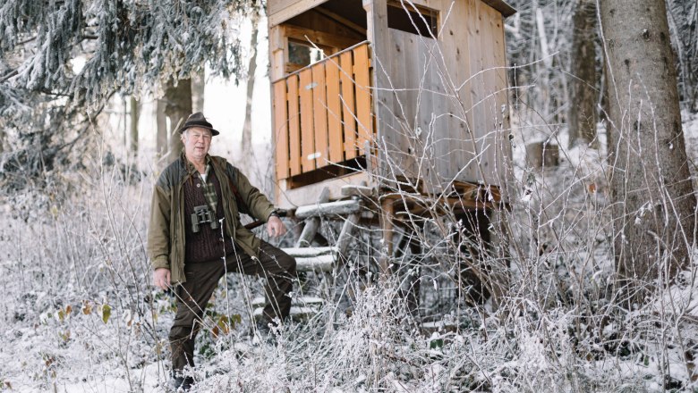 Senior boss Karl Donhauser is a passionate hunter, © Niederösterreich Werbung/ A man in hunting clothes stands next to a high seat in the snowy forest.