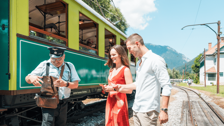 Höllentalbahn, © Österreich Werbung/Stefan Strasser A conductor in uniform shows a couple in front of a green train something from his pocket. Tracks and mountains can be seen in the background.