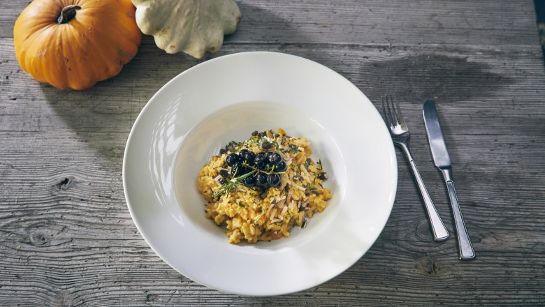 Kürbisrisotto, © Niederösterreich Werbung/Andreas Hofer Ein Teller Kürbisrisotto mit Kräutern und Beeren, daneben zwei Kürbisse auf einem Holztisch.