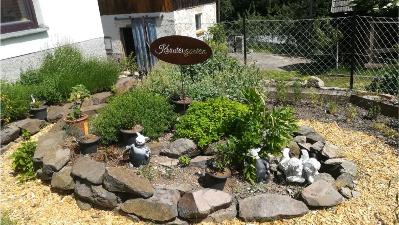 Naturgartenparadies Beisteiner - Herb garden, © Wiener Alpen A herb garden with a stone border, plants and decorations, including garden figures. A sign with the inscription 'Herb garden' is visible.