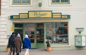 Winter, Wiener Alpen in Niederösterreich, Konditorei Nöbauer, Reichenau an der Rax, © Niederösterreich Werbung/friendship.is/Ian Ehm In der winterlichen Atmosphäre schlendern Besucher entspannt zur charmanten Konditorei, die mit ihren einladenden Fenstern und dem warmen Licht einladend wirkt. Der Duft frisch gebackener Leckereien zieht durch die kalte Luft und verspricht süße Genüsse, während die schneebedeckten Berge im Hintergrund eine malerische Kulisse bieten.