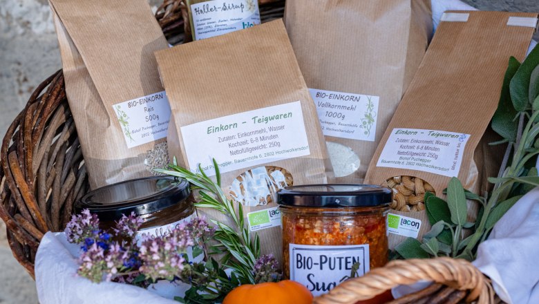 Biohof Puchegger, © Biohof Puchegger/Martina Siebenhandl Ein Geschenkkorb mit verschiedenen Produkten wie Einkornwaren, Bio-Puten Sugo und Hollersaft, dekoriert mit Kräutern und Blumen.