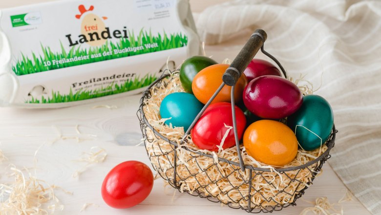 Colorful Easter eggs, © Vikografik/ Viktoria Kornfeld A basket filled with straw and colorful Easter eggs on a wooden table. Next to it, an egg carton, a red egg and a gray cloth.