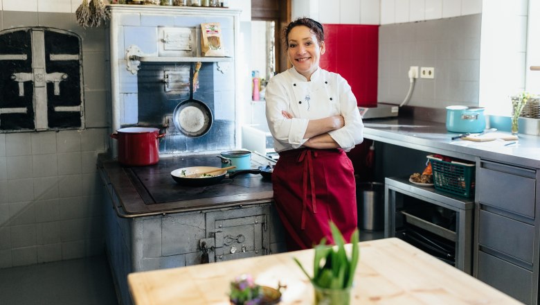 Herbalist Gerda Stocker next to her almost 100-year-old wood-fired oven, © Nicole Heiling A cook in a white jacket and red apron stands in a kitchen in front of an old stove.
