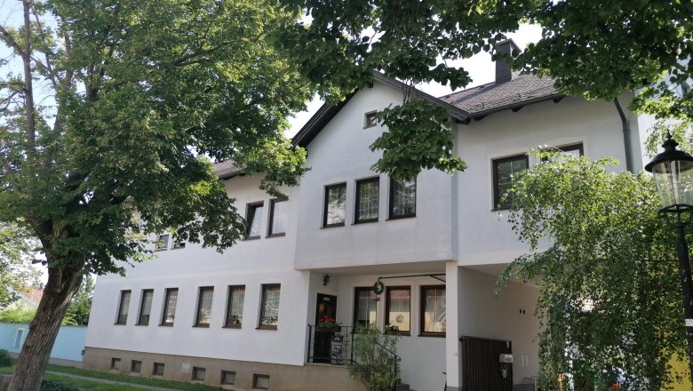 Heurigen Leeb, © Wiener Alpen Ein weißes Gebäude mit mehreren Fenstern, umgeben von Bäumen und einem Laternenpfahl.