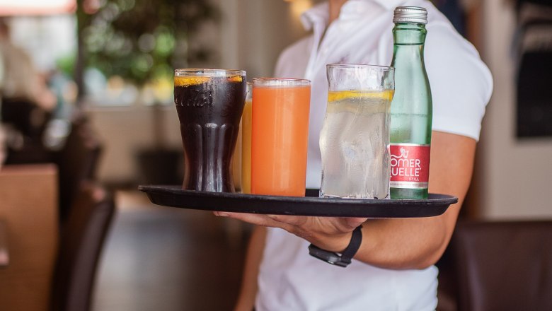 Cozy ambience., © Koll GmbH A smiling waitress in a restaurant carries a tray with various drinks.