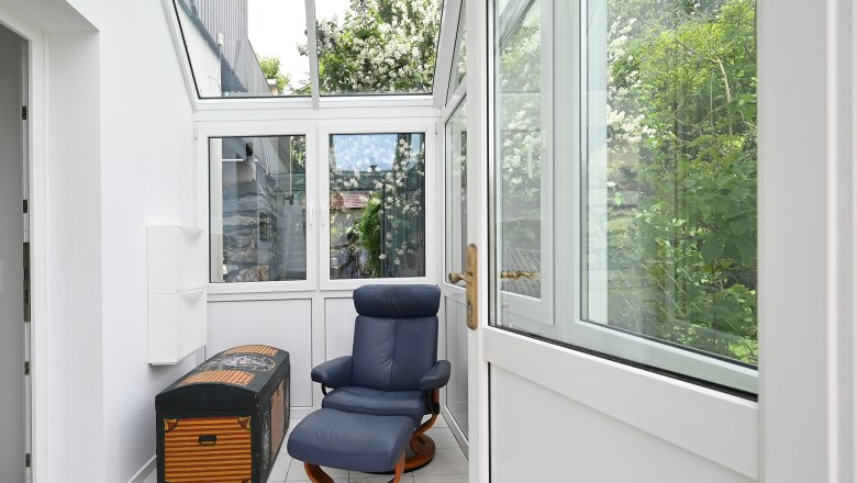 Vorraum, © Christine Kuppelwieser Ein heller Vorraum mit einem blauen Sessel und einer dekorativen Truhe, umgeben von großen Fenstern mit Blick auf grüne Pflanzen.