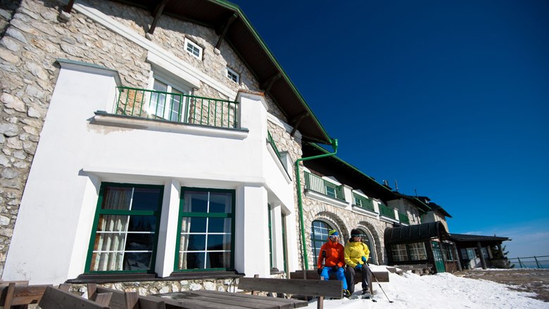 Mountain inn Raxalpe in winter, © Wiener Alpen - Ziegler Two people in winter clothing in front of a mountain inn with snow-covered ground and blue sky.