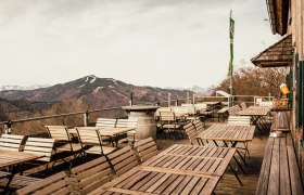 Von der Terrasse, © Stefanie Rysavi Leere Terrasse mit Holztischen und -stühlen, Berglandschaft im Hintergrund.