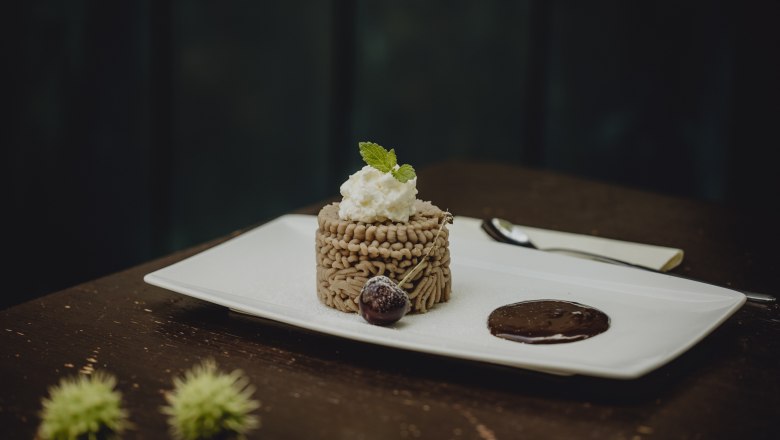 Ganzjähriger Favorit: Kastanienmousse, © Niederösterreich-Werbung/Sophie Menegaldo Ein Dessert aus Kastanienmousse auf einem weißen Teller, garniert mit Sahne und Minzblatt, daneben eine Schokoladensauce und eine Kirsche.