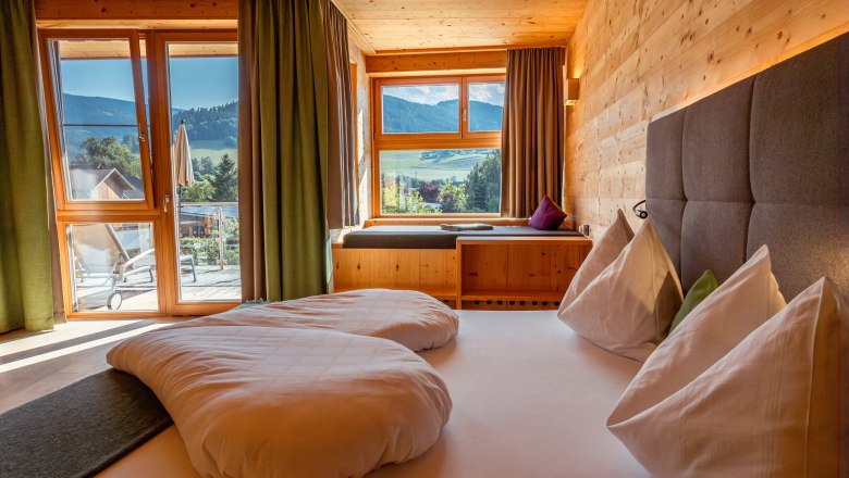 Naturhotel Molzbachhof, © Niederösterreich Werbung / Maximilian Pawlikowsky Innenansicht eines Hotelzimmers mit Holzwänden, großem Bett und Blick auf die Berge durch ein Fenster.