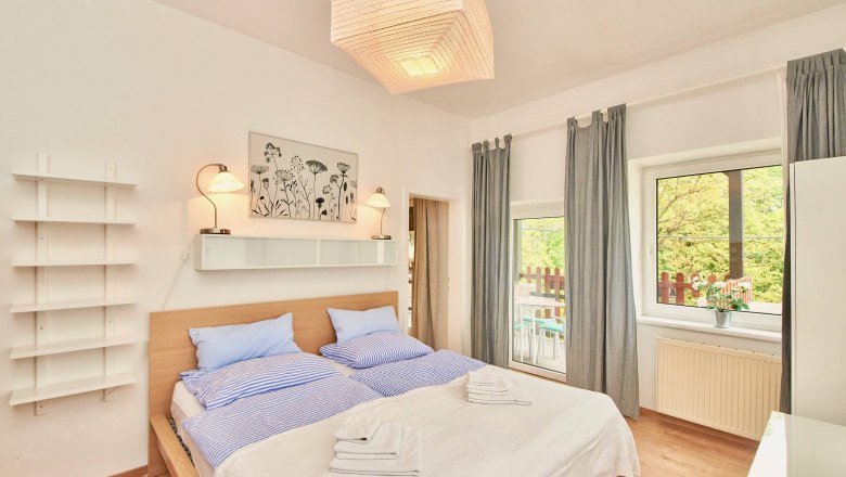 Top 2 - Schlafzimmer, © VP FeWo OG Helles Schlafzimmer mit Doppelbett, blauer Bettwäsche, zwei Lampen, einem Bild an der Wand und einem Fenster mit Blick auf einen Balkon.