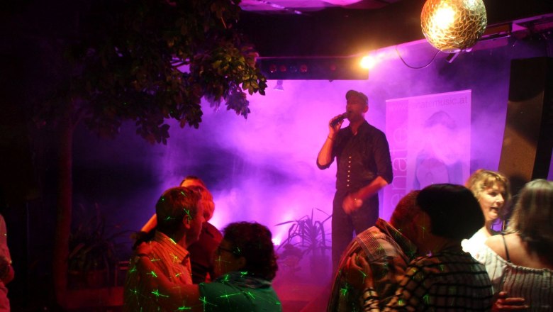 Livemusik und Tanzabende im Preiner Stadl, © Scharfegger’s Raxalpen Resort Ein Sänger auf einer Bühne mit Mikrofon, umgeben von tanzenden Paaren und bunten Lichtern.