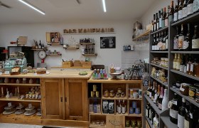 "Das Kaufhaus" in Aspang, © Das Kaufhaus, Peter Wochesländer Innenansicht eines kleinen, gemütlichen Ladens mit Holzregalen, gefüllt mit Weinflaschen, Lebensmitteln und einer alten Registrierkasse.